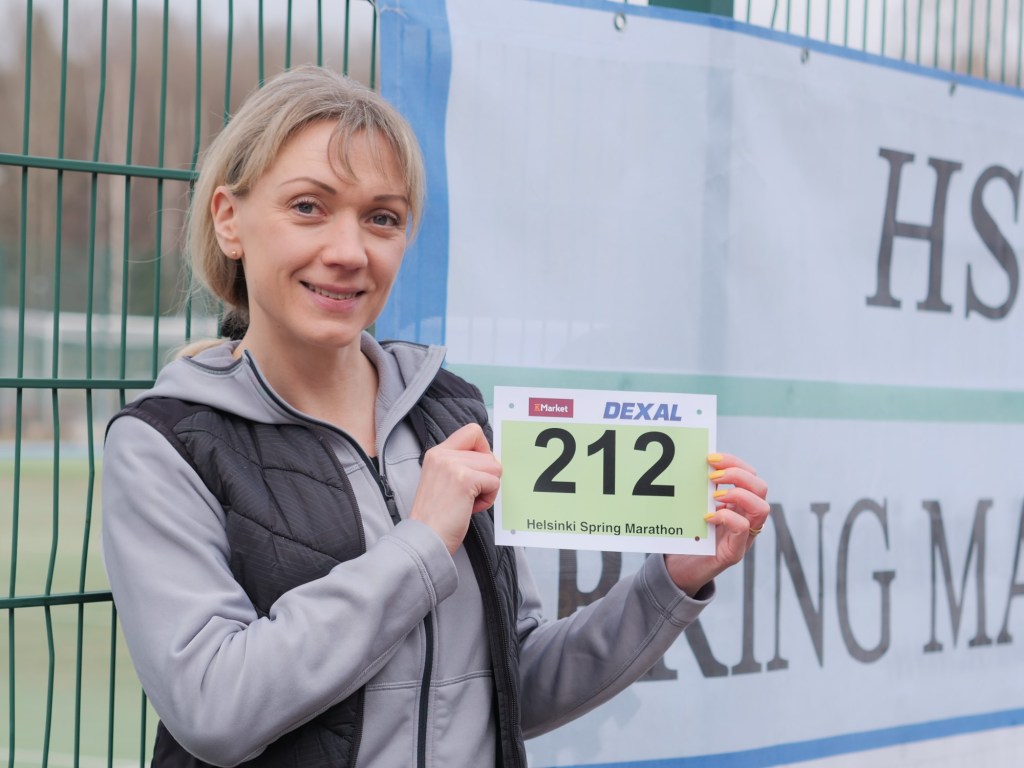 nainen seisoo numerolappu kadessaan mainosjulisteen edessa