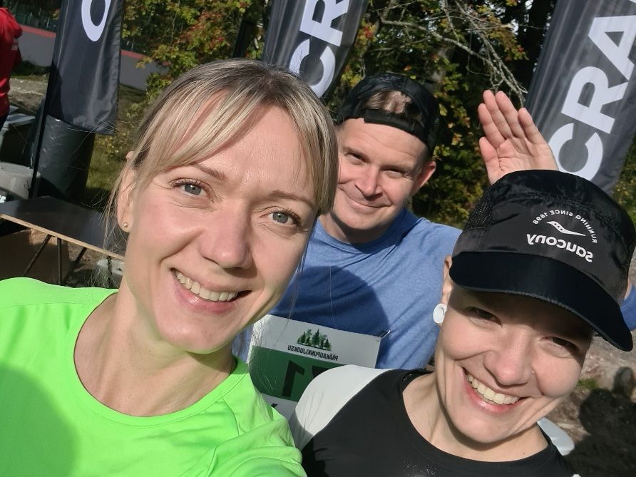 three runners smiling at the camera and a man waving on the background